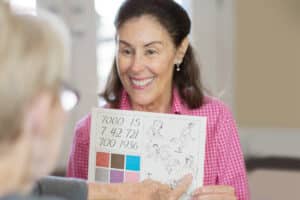 Smiling brown haired woman in pink check shirt showing a person pictures and the person pointing to one of the pictures.