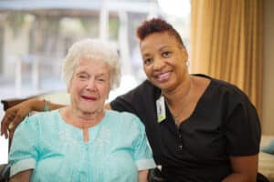 Medical professional in black scrubs with her arm around elderly woman in light blue blouse.