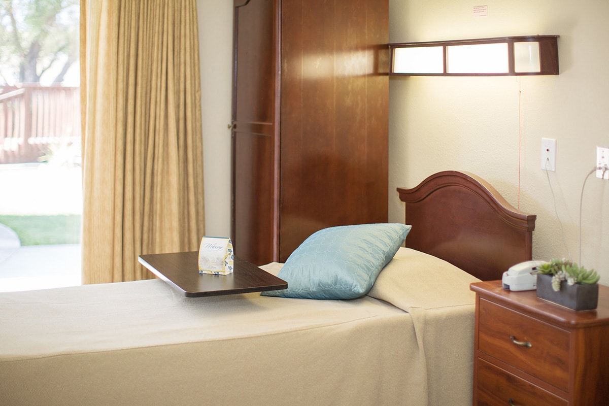 Patient room with bed made, night stand, curtains and blue decorative pillow with view of courtyard.