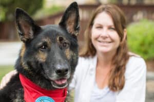 German shepherd dog with red bandana smiling at camera with woman with long brown hair smiling in background.