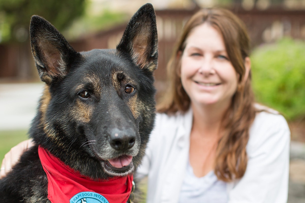 German shepherd dog with red bandana smiling at camera with woman with long brown hair smiling in background.